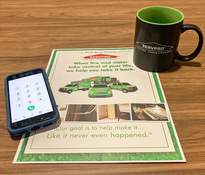 Flyer about SERVPRO laying on a wooden desk with a black SERVPRO mug and phone with the number dial showing on screen